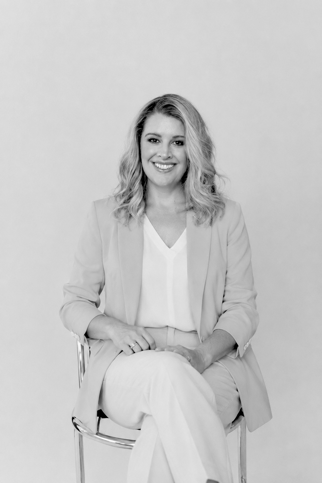 Black and white portrait of a smiling young woman with wavy hair, wearing a blazer and sitting on a chair against a plain background.