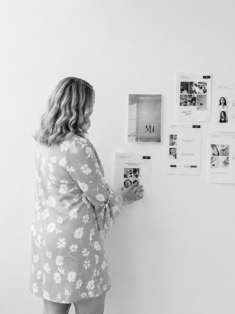 Woman in a floral dress looking at a wall with papers and photos attached.