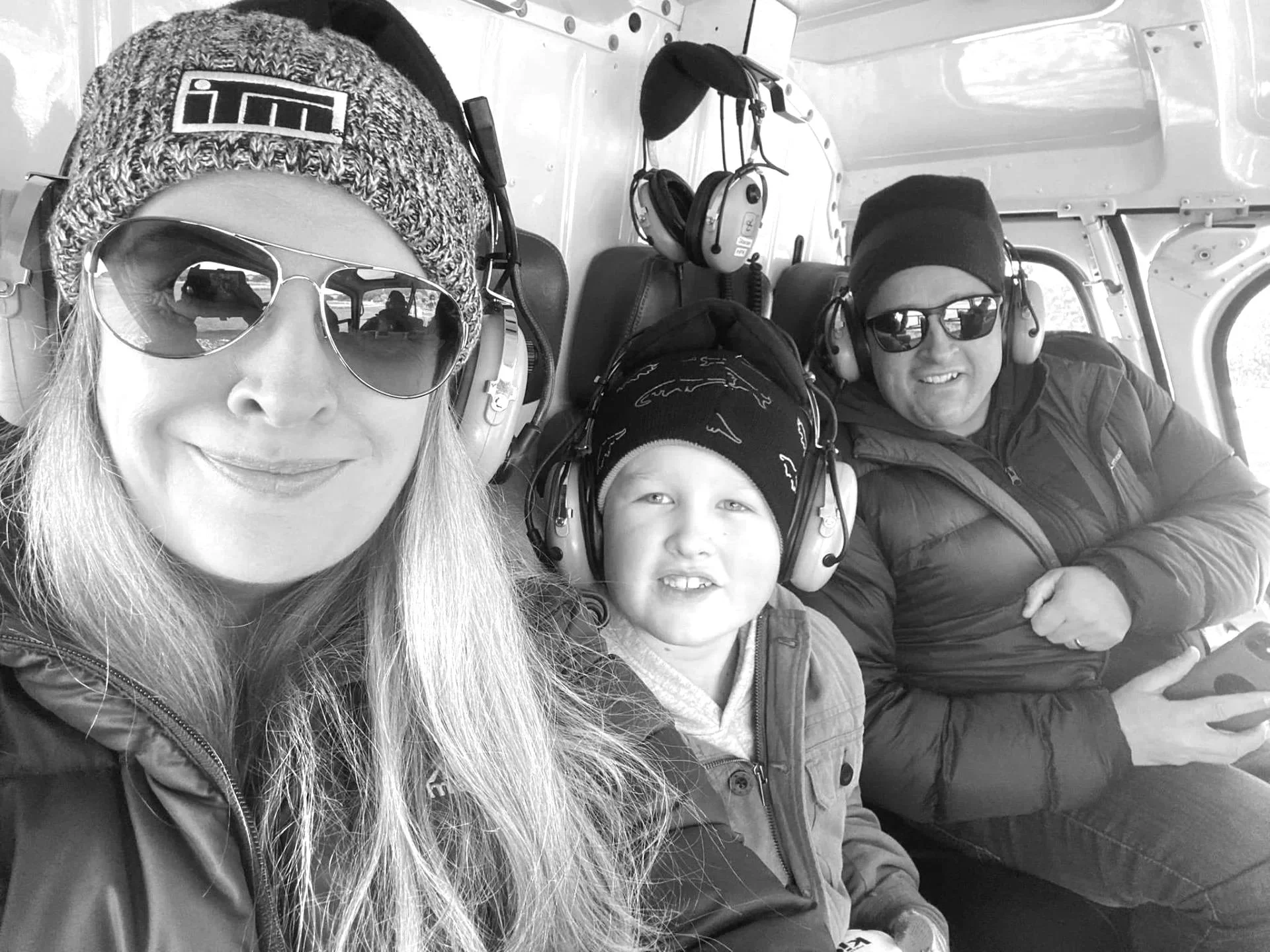 A woman, a young boy, and a man sitting inside an aircraft, all wearing headphones and sunglasses. The woman is wearing a knitted hat and the man is wearing a beanie. The woman is taking the selfie, and they are smiling.