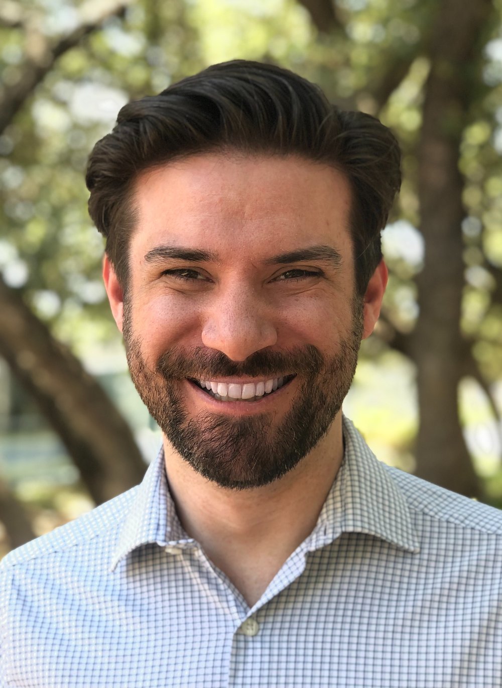 A man with dark hair and a beard smiling outdoors with trees in the background.
