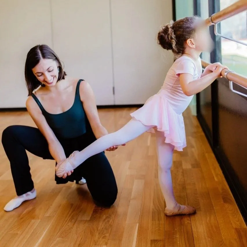 A young girl in a pink ballet dress stretching with her leg on a ballet barre, while an adult woman in ballet attire helps her stretch, both inside a dance studio with wooden floors and large windows.
