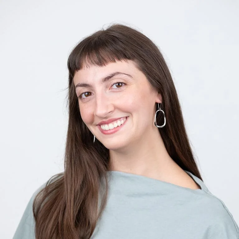 A woman smiling with long brown hair, wearing a light gray top and silver earrings, against a plain white background.