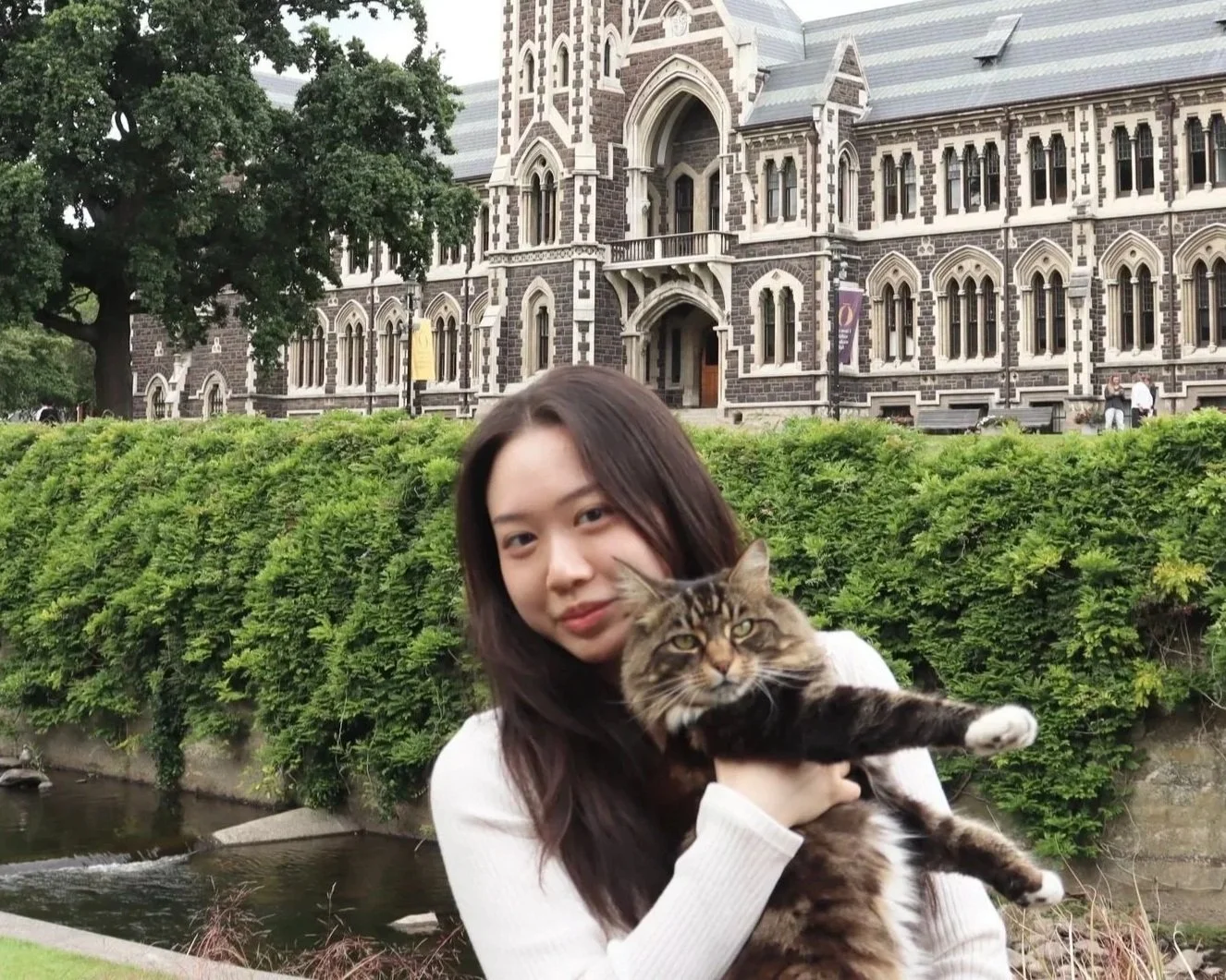 A young woman holding a large tabby cat outdoors in front of a historic building with gothic architecture and lush green trees.