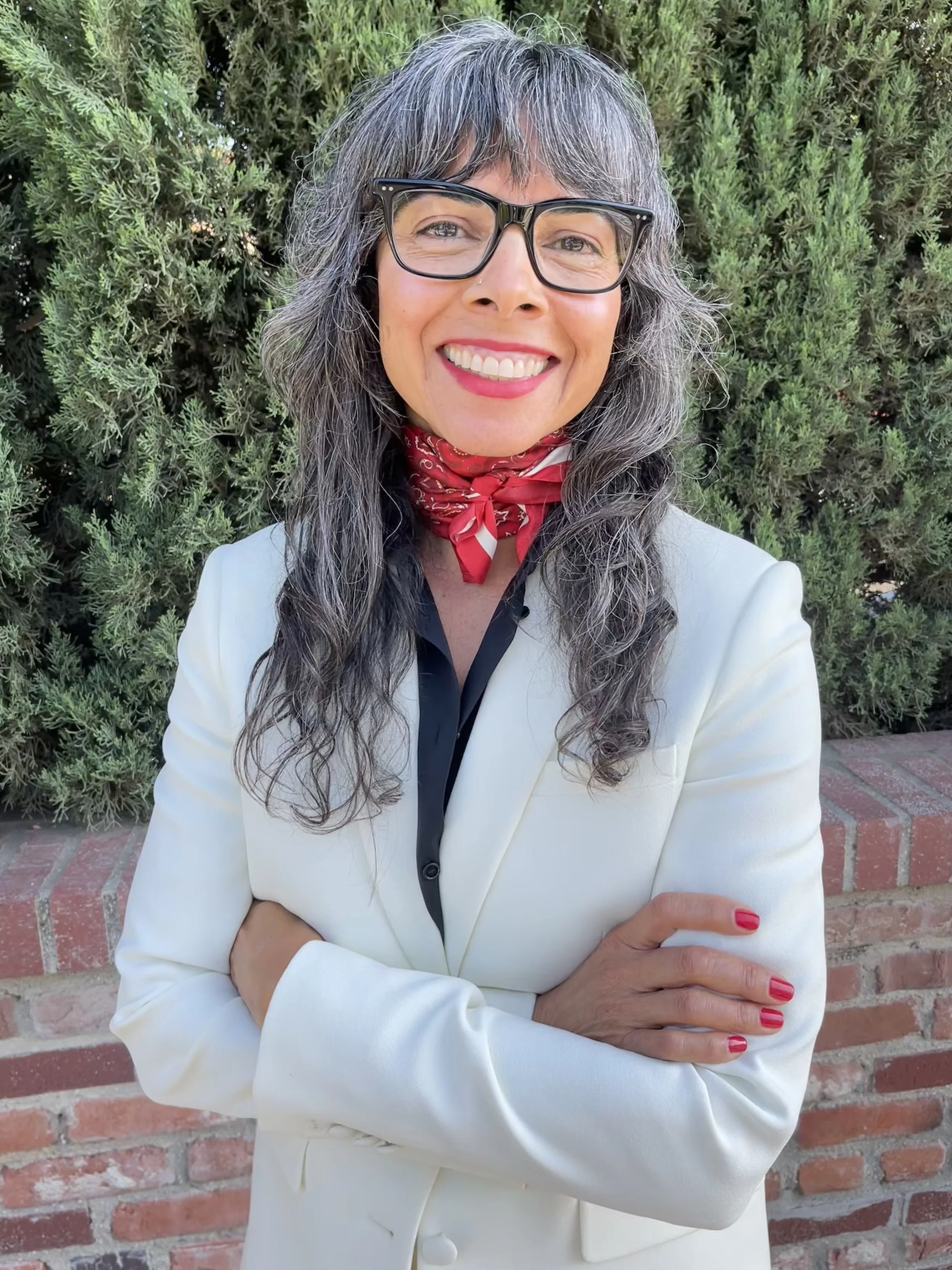 Nancy Elliott smiling outdoors in a cream blazer and red neckerchief, arms crossed, in front of greenery