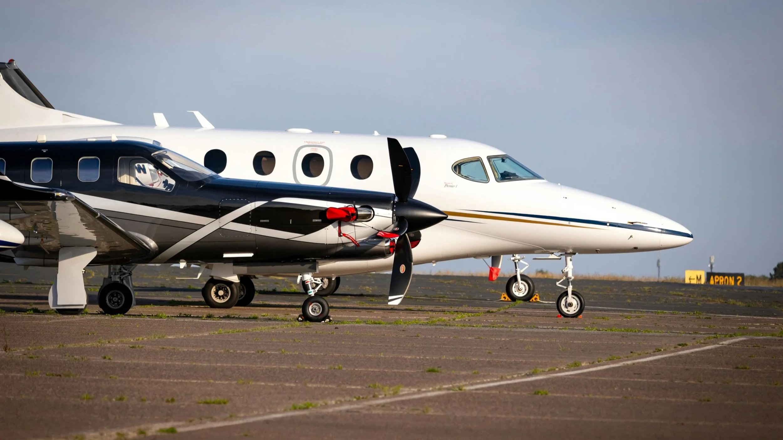 View of two private jets parked on an airport tarmac during daytime, with a clear sky in the background.