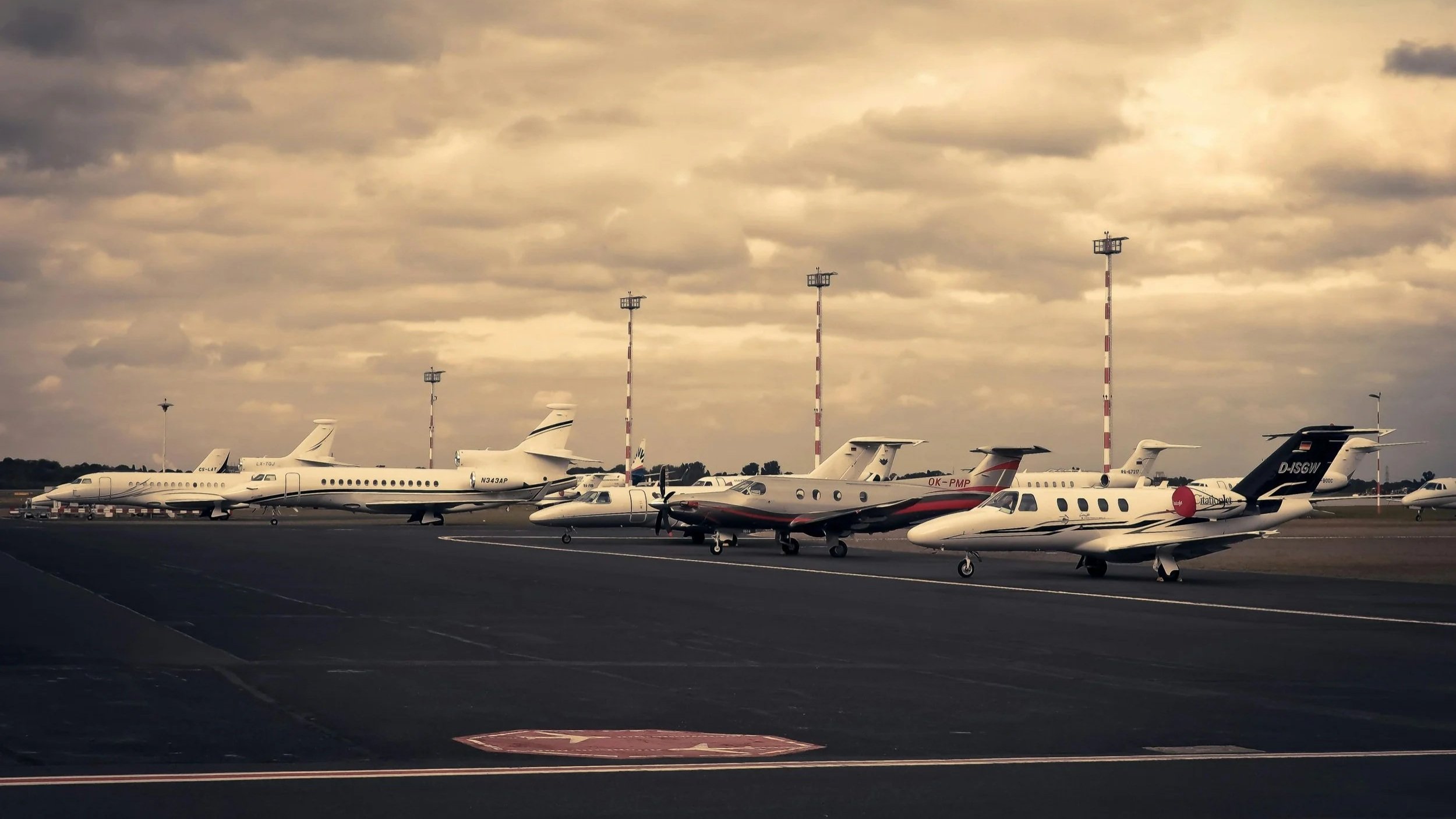 Multiple private jets parked on an airport tarmac under cloudy sky, with tall light poles in the background.