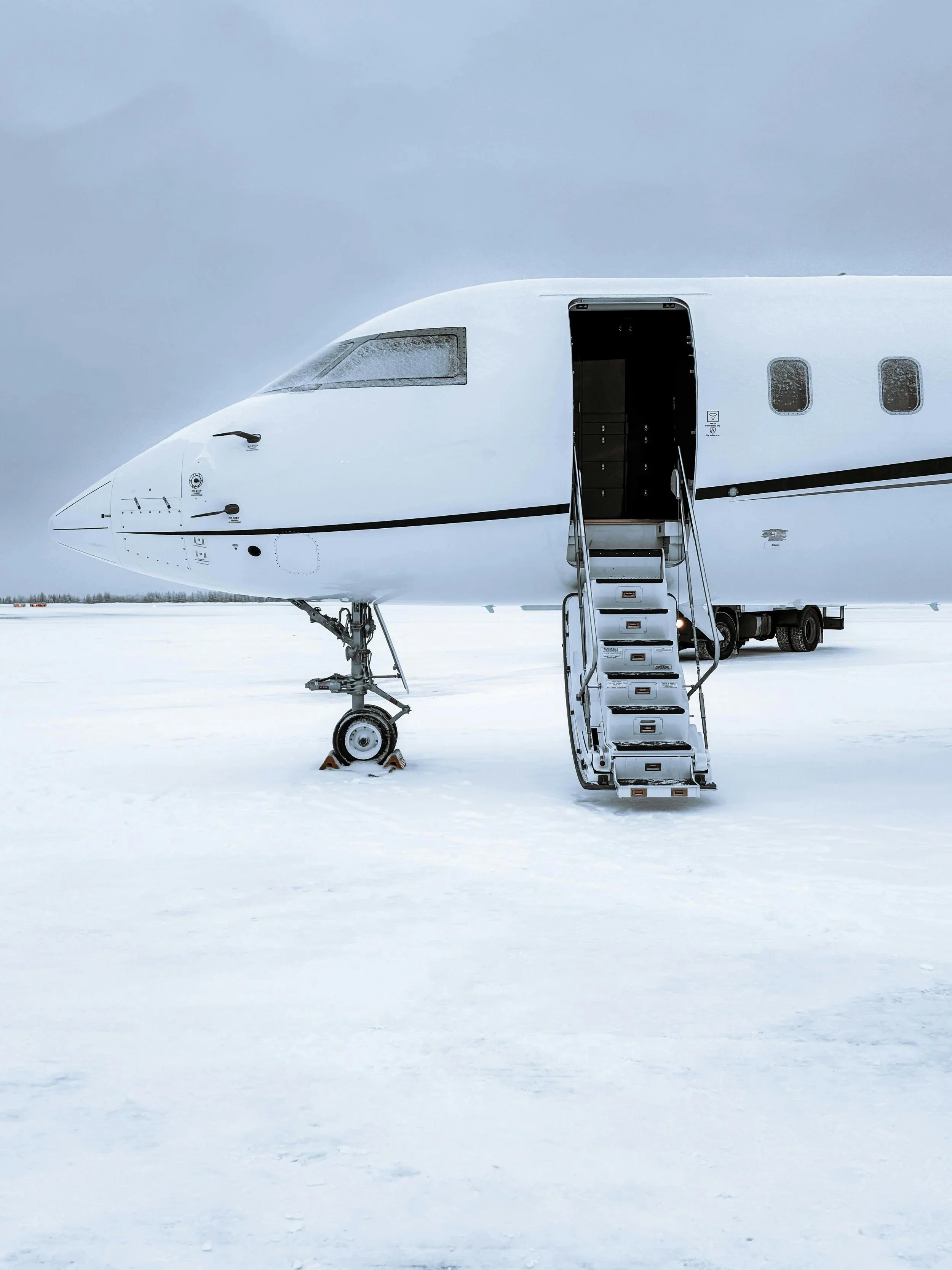 A white airplane parked on snowy ground with stairs leading up to the open door, under a cloudy sky.