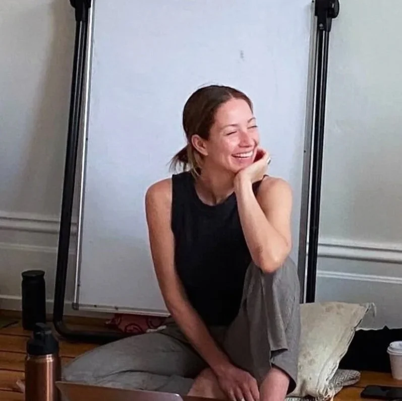 A woman sitting on a pillow on the floor, smiling and laughing, with her hand resting on her face, in a room with a whiteboard and some equipment in the background.