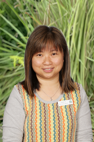 A woman with straight brown hair wearing a colorful patterned top and a silver cross necklace, standing in front of tall green grass.