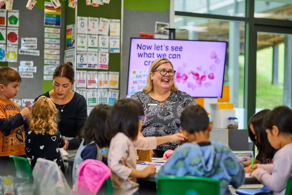 A classroom scene with children and teachers, a woman smiling in front of a purple screen that says 'Now let's see what we can create'. The classroom has educational posters and a window showing greenery outside.
