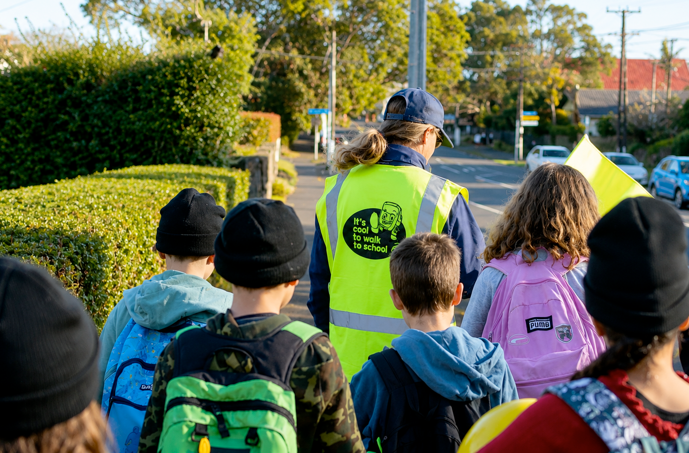 A group of children with backpacks walking on a sidewalk with a crossing guard wearing a high-visibility vest guiding them across the street.
