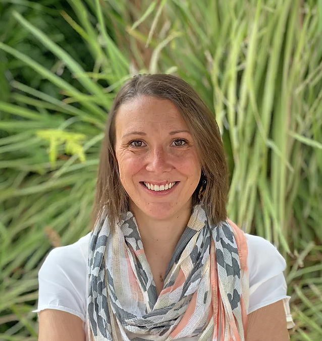 A woman with brown hair smiles at the camera, wearing a white shirt and a patterned scarf, with tall green plants in the background.
