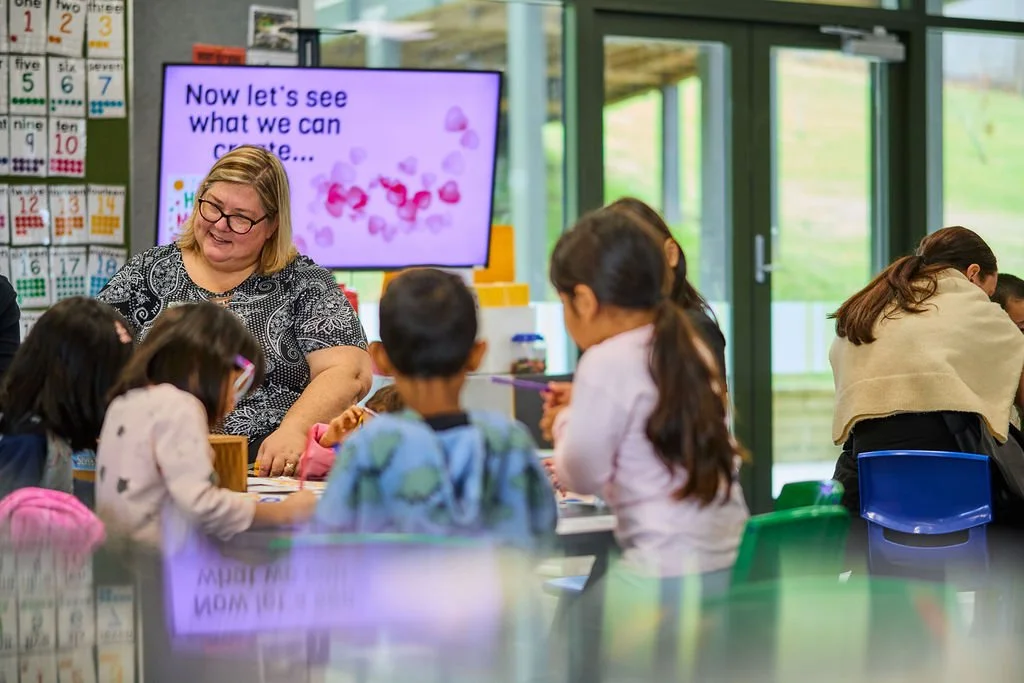 A teacher smiling and interacting with students in a classroom with colorful chairs and educational posters, a digital screen displaying a message, and large windows letting in natural light.