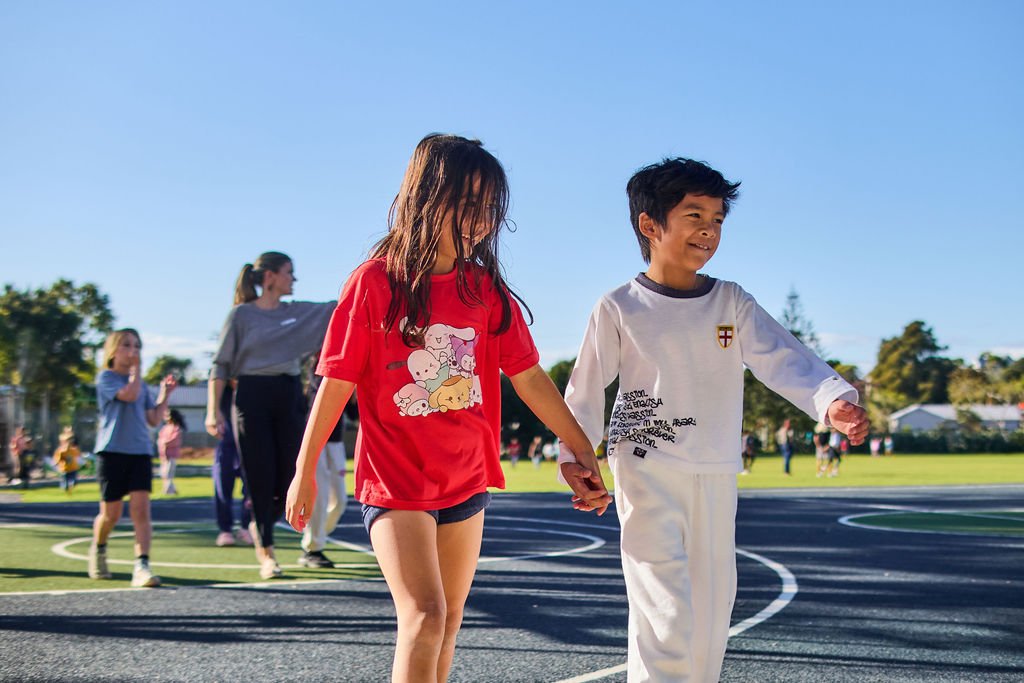 Two children walking hand in hand on a sports court, with other children and adults in the background, under a clear blue sky.