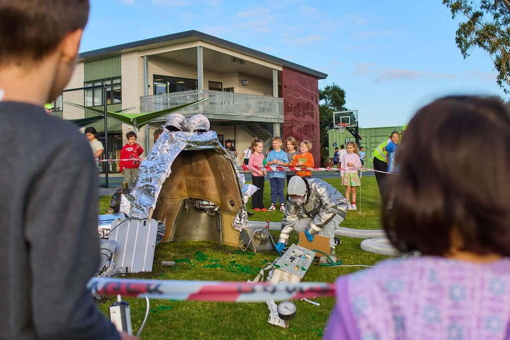 Children gathered outdoors watching a science demonstration involving a homemade rocket or volcano explosion with a person in a protective suit working on the setup.
