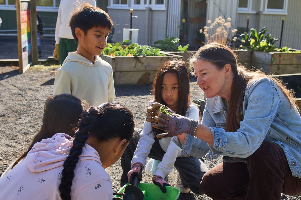 Adult woman in denim jacket showing freshly dug plant roots to children in a garden