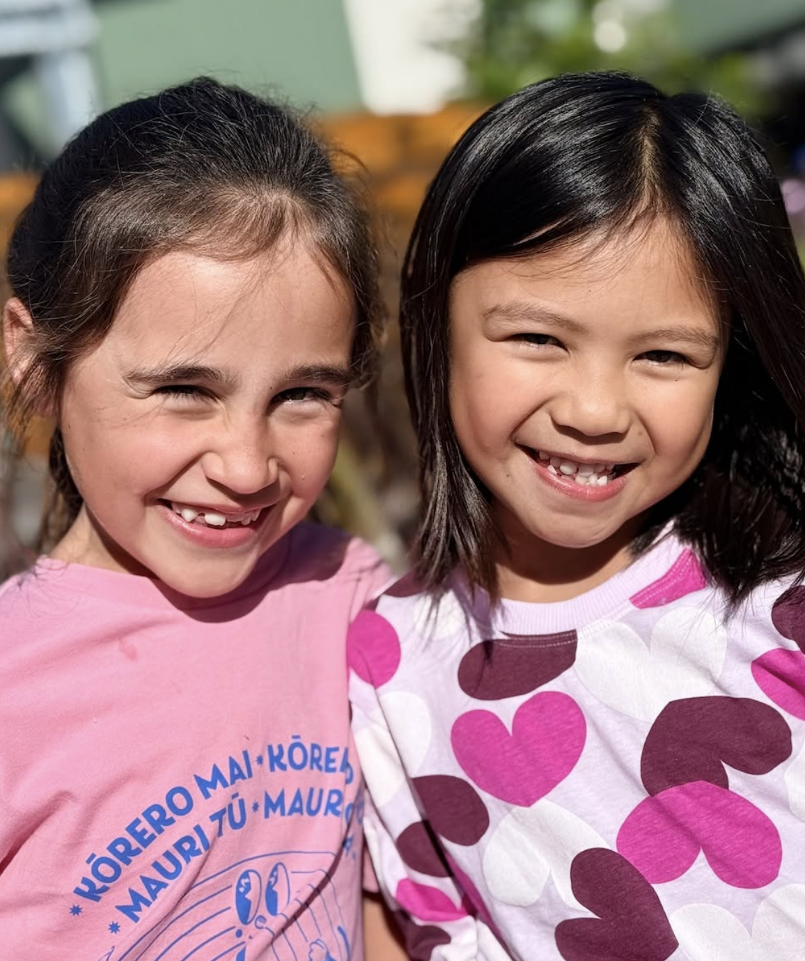 Two smiling young girls standing close together outdoors, one with dark hair and the other with brown hair, both wearing colorful shirts with heart and dot patterns, bright sunlight, green background.