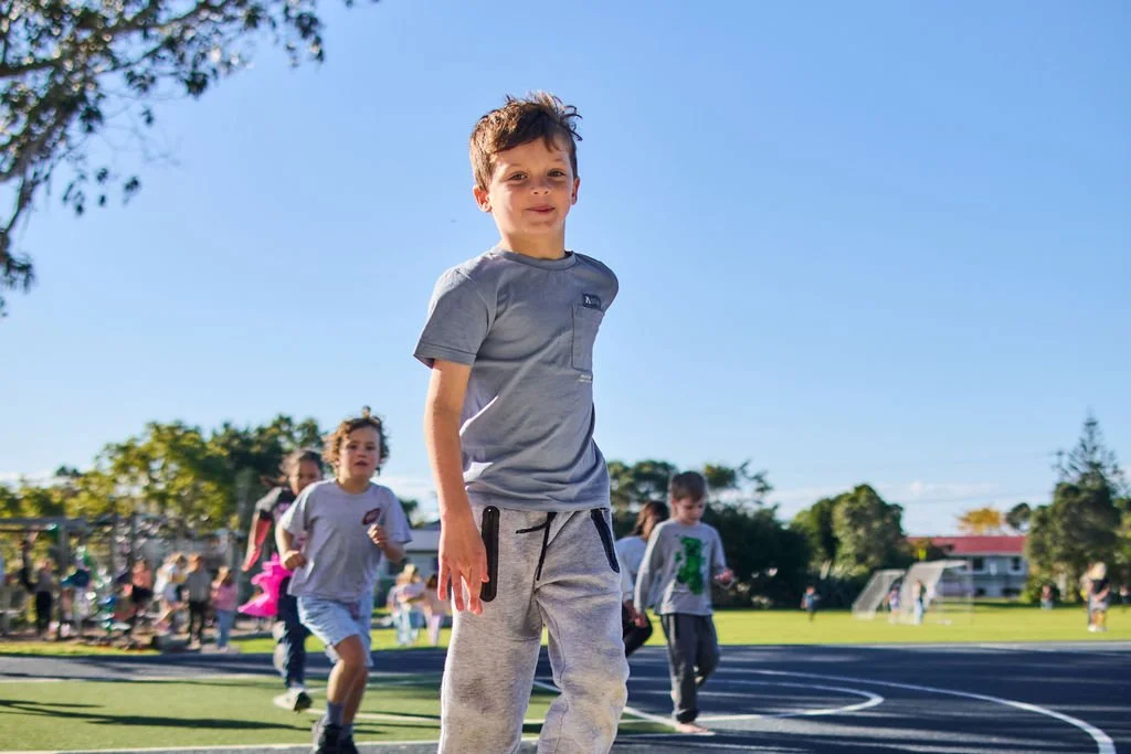 A young boy in a gray T-shirt and sweatpants standing on a school playground during recess, with other children playing and running in the background under a bright blue sky.