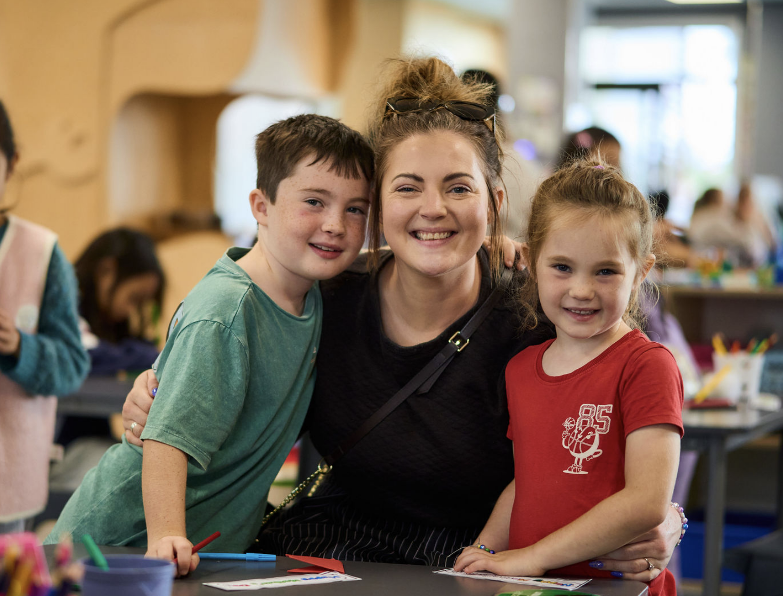 A woman and two children smiling and posing together in a classroom setting, with other children and arts and crafts supplies visible in the background.