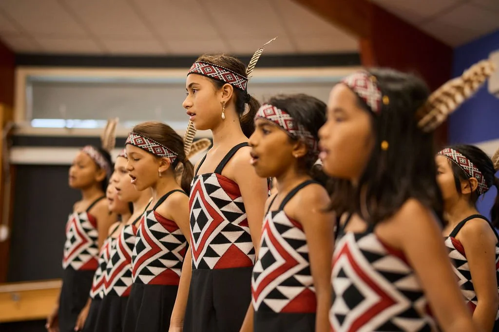 Group of young girls dressed in traditional Indigenous dance attire with geometric patterns, standing in a line during a performance.