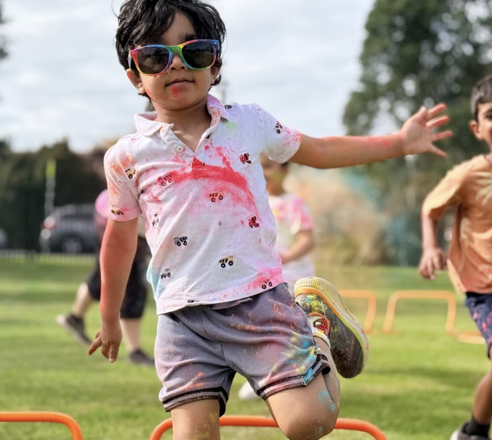 Child wearing rainbow-colored sunglasses and a white shirt with colorful splashes of paint, actively participating in an outdoor youth sports activity on a grassy field, with other children in the background.