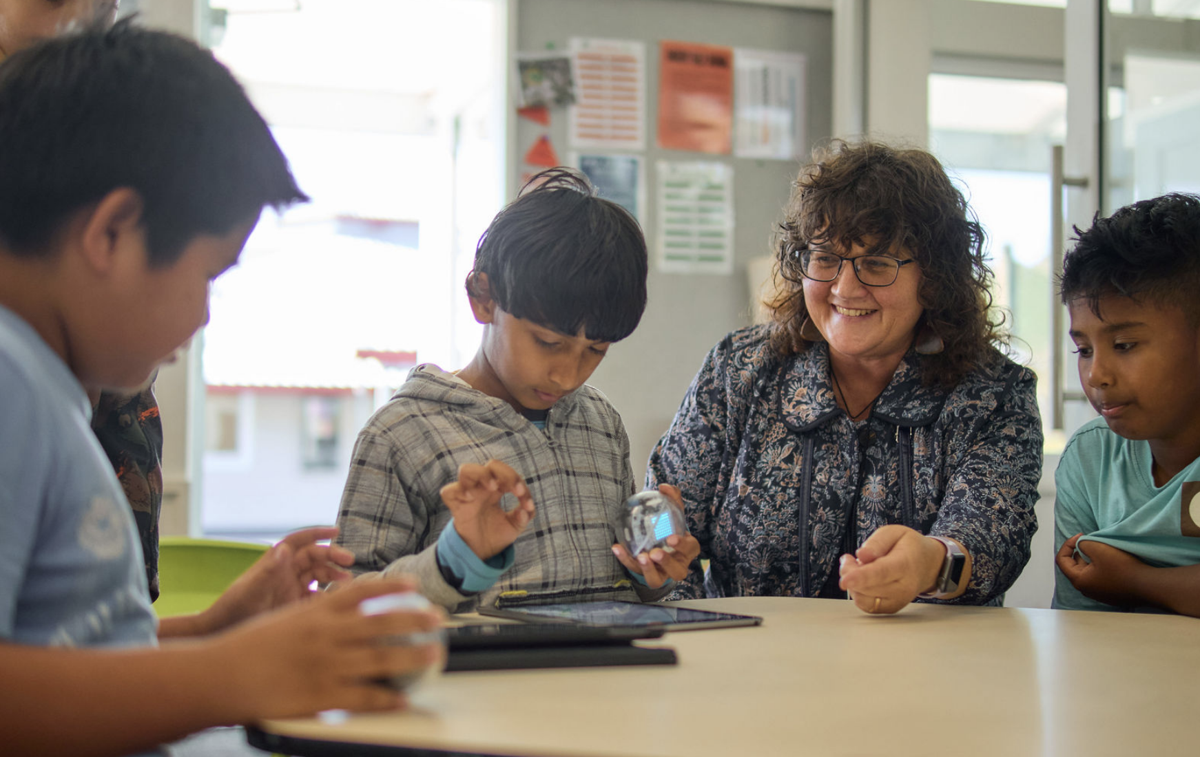 A diverse group of children and an adult woman gathered around a table, looking at a small robot in a classroom setting.