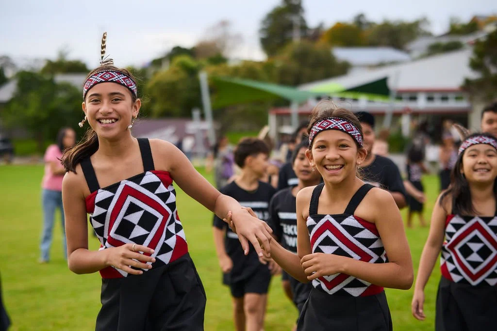 Two young girls wearing traditional Maori attire, including headbands and patterned tops, holding hands and smiling during a cultural event outdoors with other people in the background.