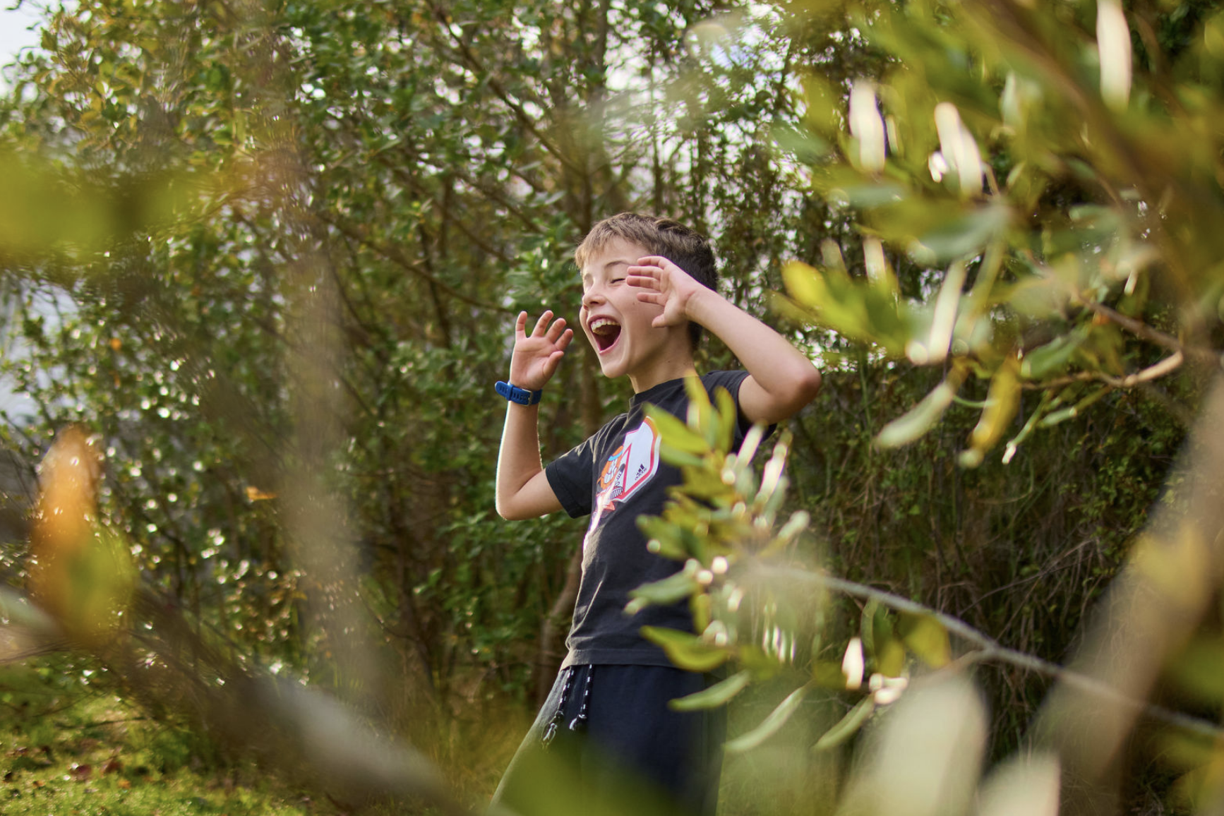 A young boy with short hair, wearing a black t-shirt and a blue wristband, smiling and making a playful gesture with his hands in a lush, green outdoor setting surrounded by trees and foliage.