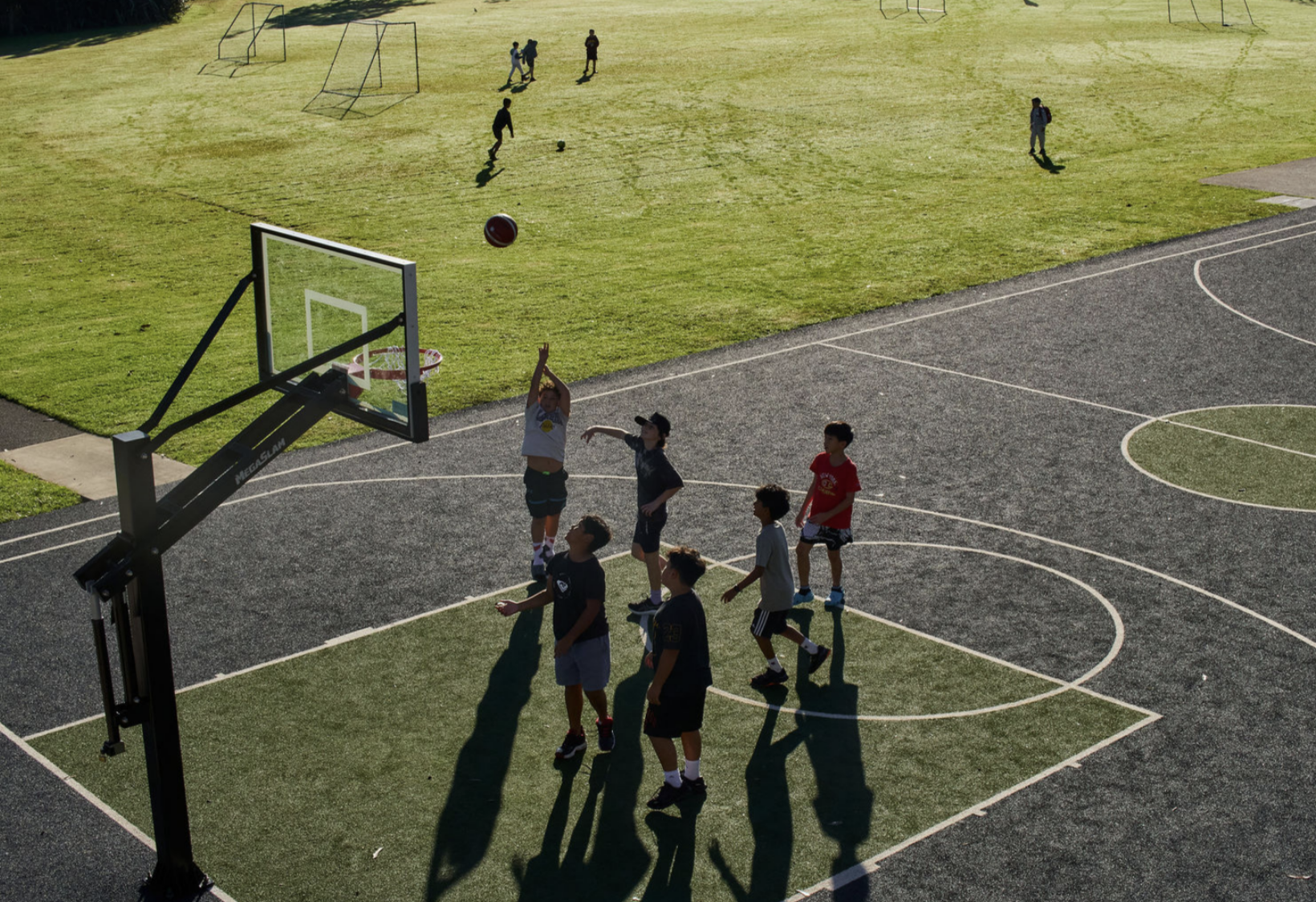 Young boys playing basketball on an outdoor court with a portable backboard, some boys are shooting and others are watching, and a group is playing soccer on the grassy field in the background.