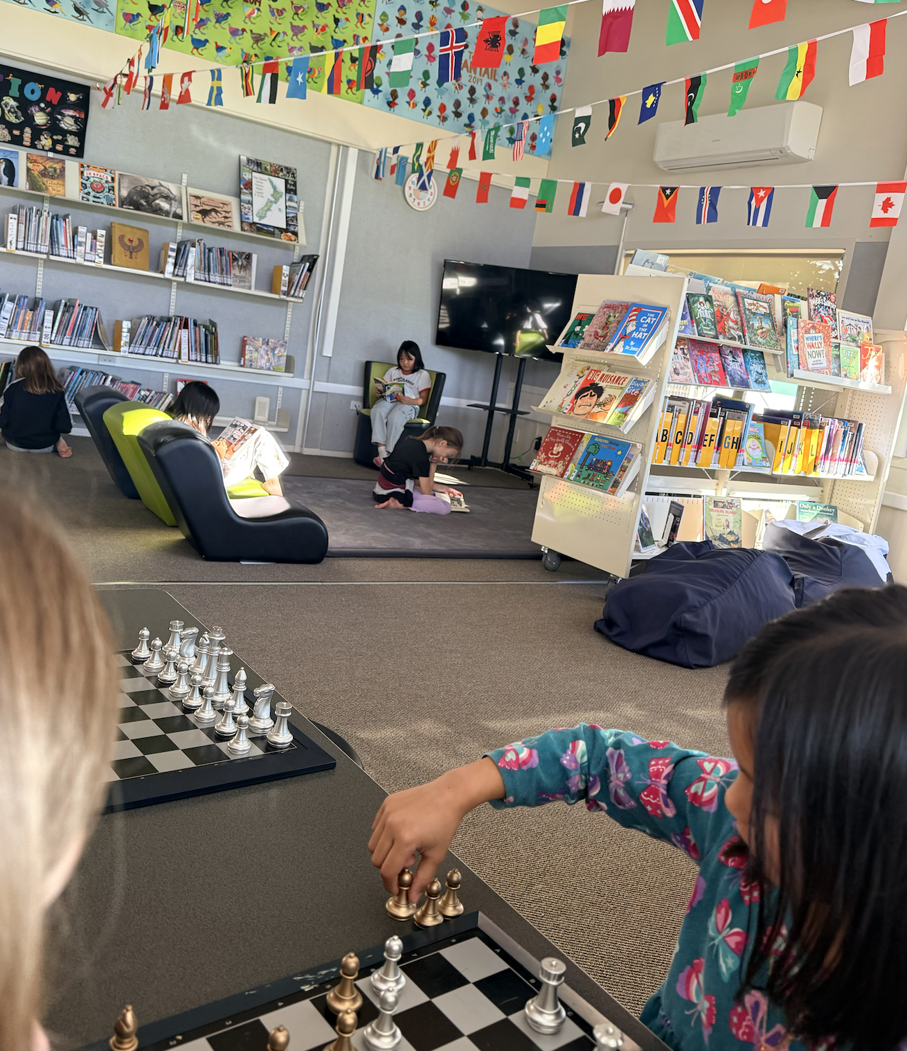 Children sitting and reading in a library with bookshelves, international flags hanging from the ceiling, a chessboard in the foreground, and a large television screen on the wall.