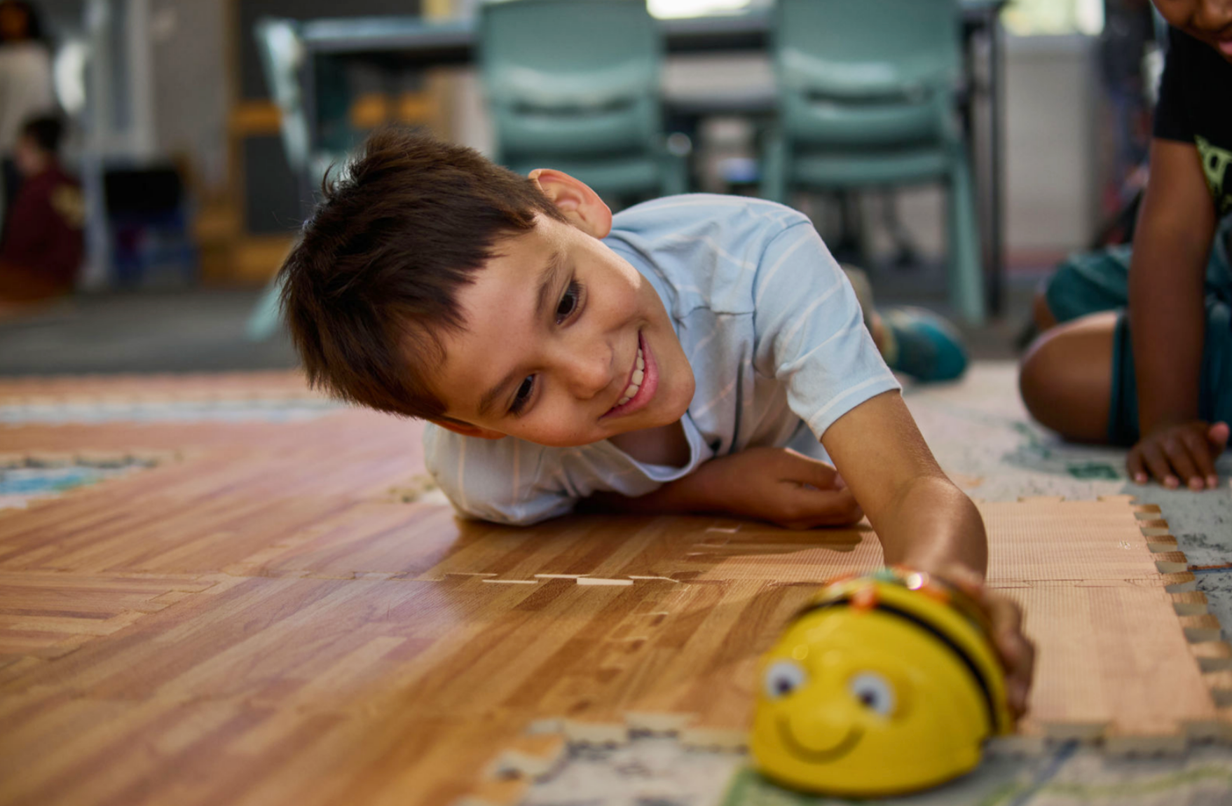 A young boy lying on the floor, reaching towards a small robot with a smile in a classroom with chairs in the background.