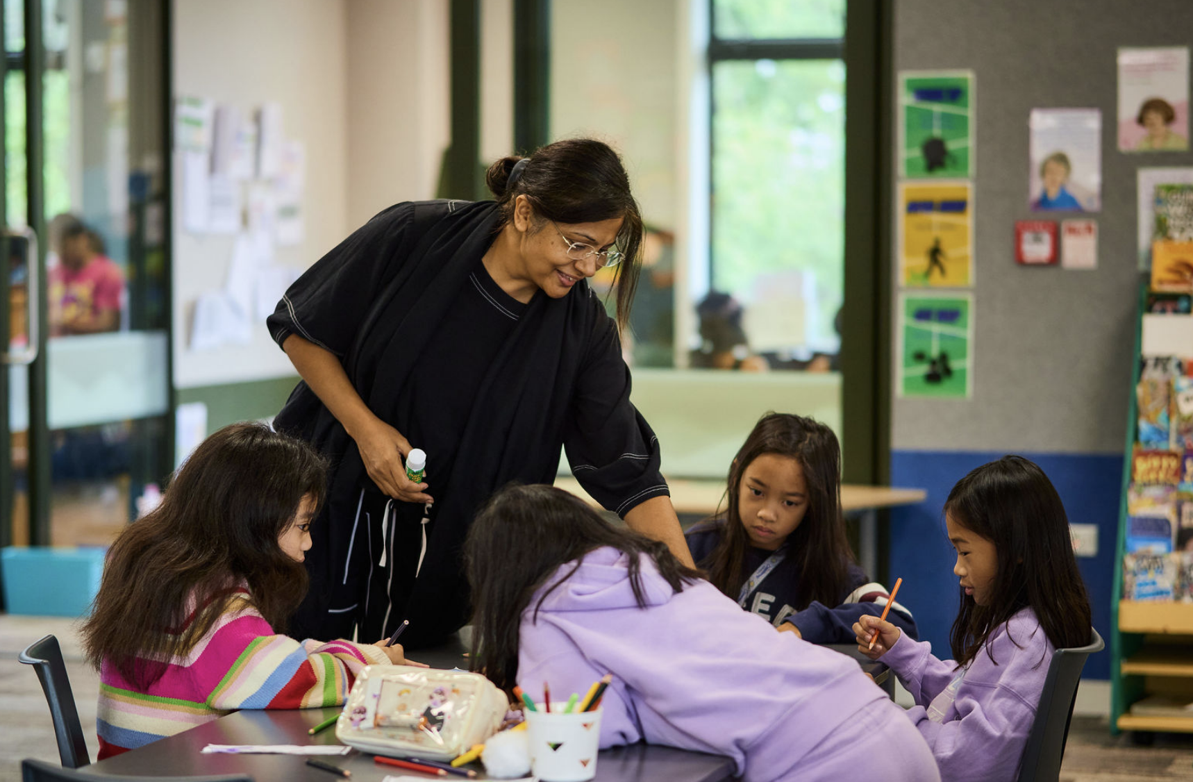 A woman with glasses and a black top helps four young girls with long hair and light to medium skin tones with their activity at a table in a classroom. The girls are focused on their work, with colored pencils and paper on the table. The classroom has colorful posters on the walls and a window showing greenery outside.