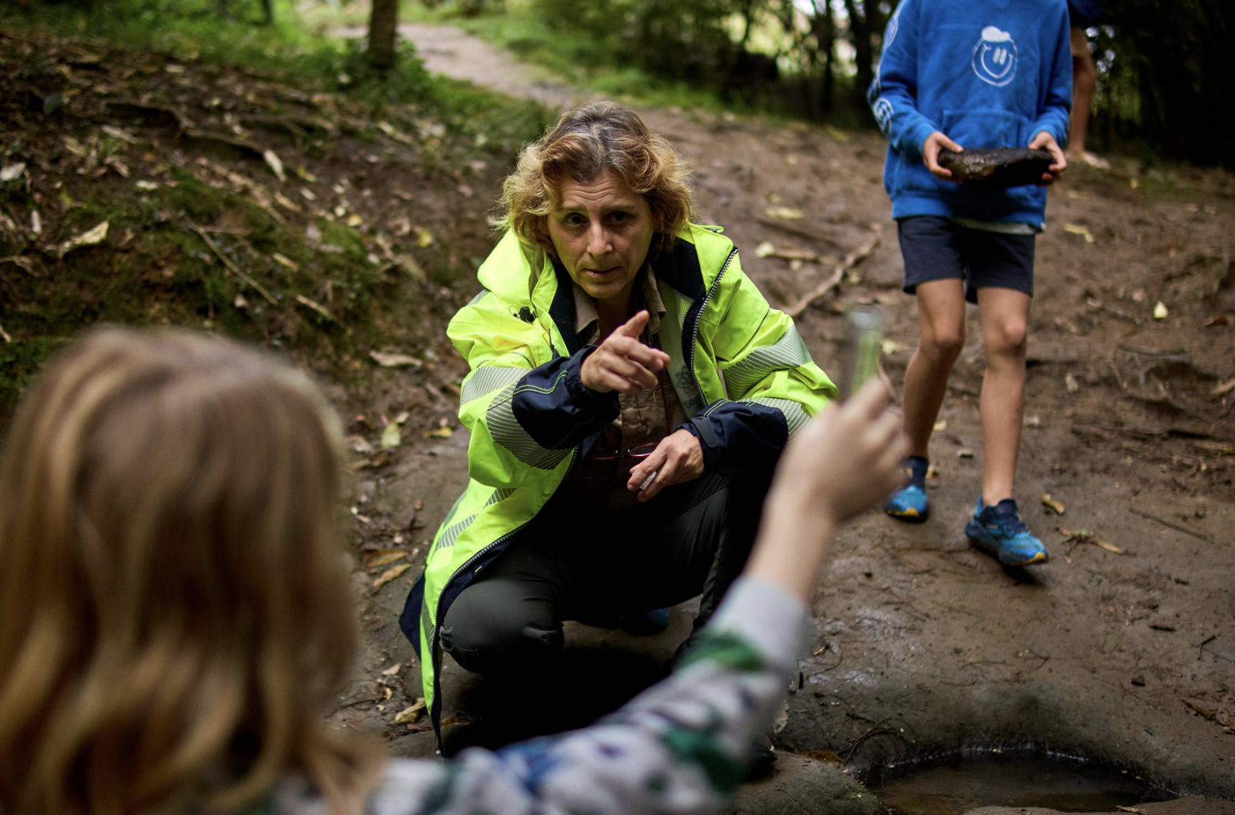 A woman in a bright yellow jacket crouches on a muddy trail in a wooded area, pointing towards something. Two children, partly out of the frame, appear to be engaged with her, one holding a small object, and another walking on the trail. The scene suggests an outdoor educational or nature exploration activity.
