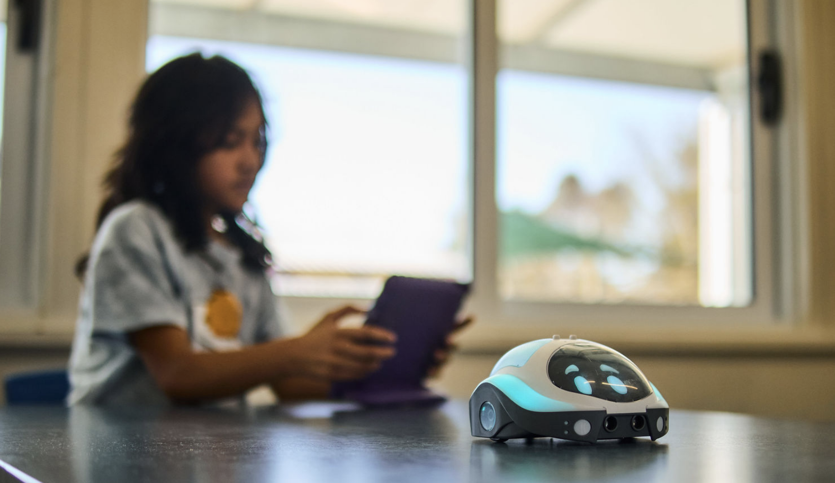 A small robot with a white and blue body, black windows, and multiple sensors sitting on a black table. In the background, a girl with long dark hair wearing a white shirt with a badge is looking at a tablet near a window.