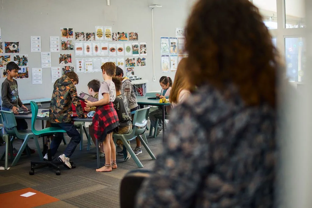 Students in a classroom working at tables with educational posters and photos on the walls, seen from behind a woman with curly hair observing.