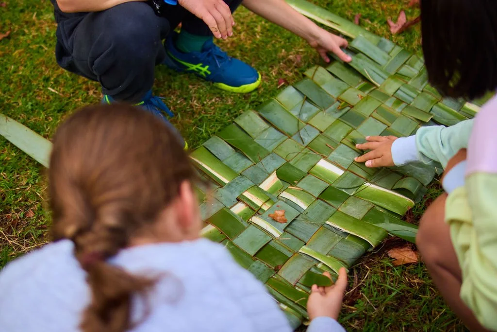 Children working together outdoors to construct something using large green leaves on the ground.