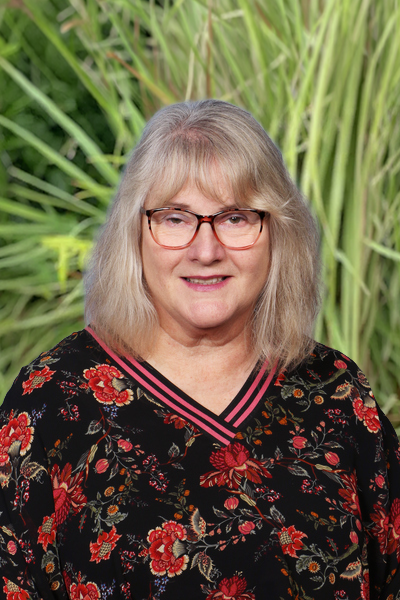 A middle-aged woman with gray hair and glasses smiling outdoors in front of tall green grass, wearing a black floral blouse.