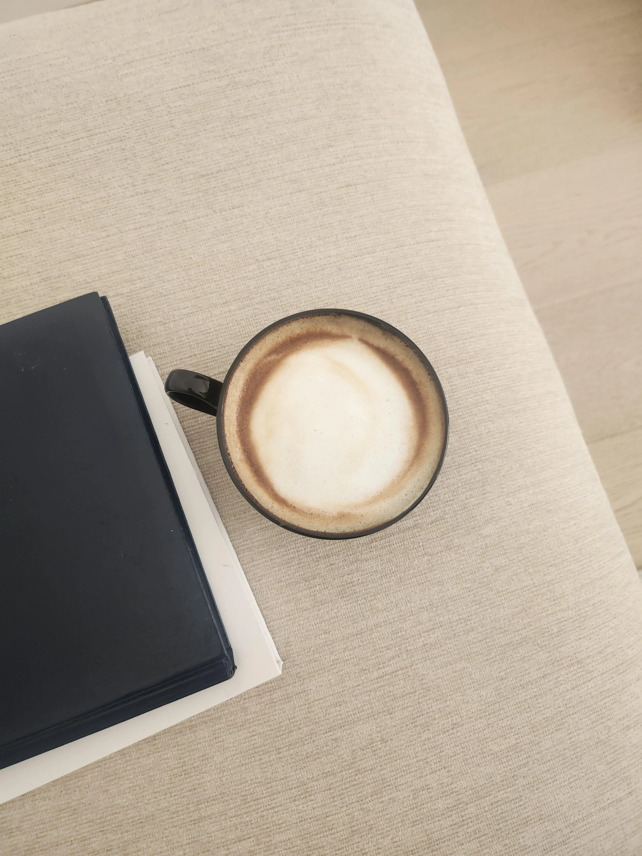A black hardcover notebook on a white tray next to a ceramic mug filled with a frothy cappuccino on a beige textured surface.