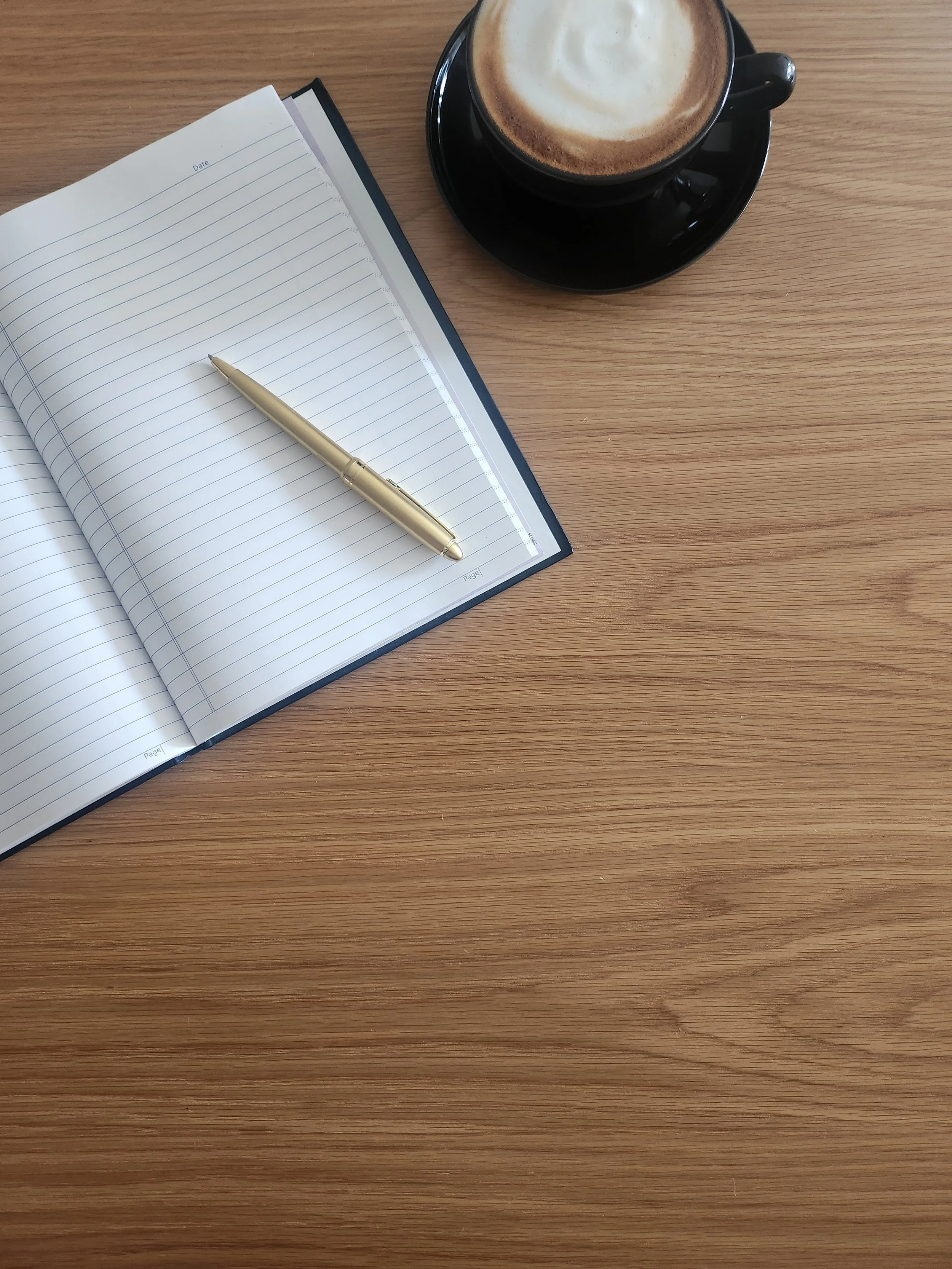 Open lined notebook with a gold pen on a wooden table, accompanied by a black cup of coffee with foam on top, placed on a saucer.