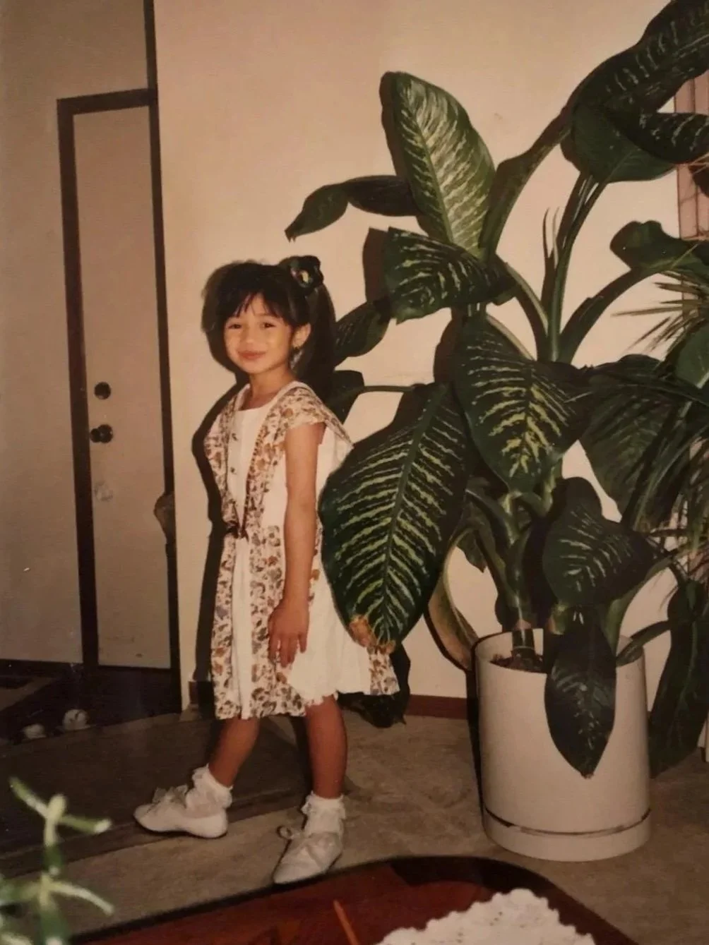 A young girl with a hairstyle, wearing a white dress with floral patterns, standing indoors next to a large leafy green houseplant in a white pot, smiling at the camera.