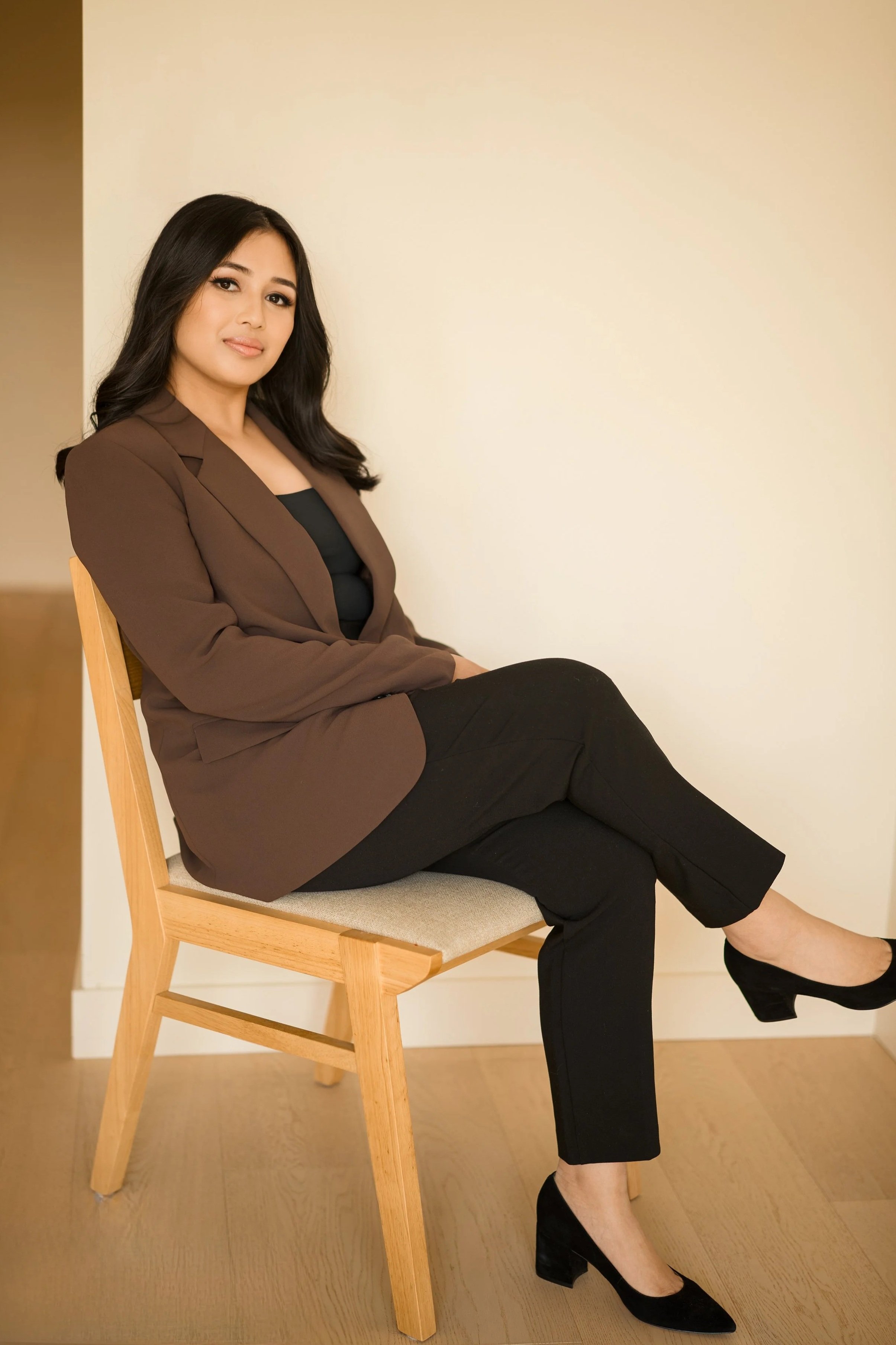 A woman sitting sideways on a wooden chair with a beige cushion, wearing a brown blazer, black top, black pants, and black heeled shoes, with a neutral expression, sitting against a plain light-colored wall.