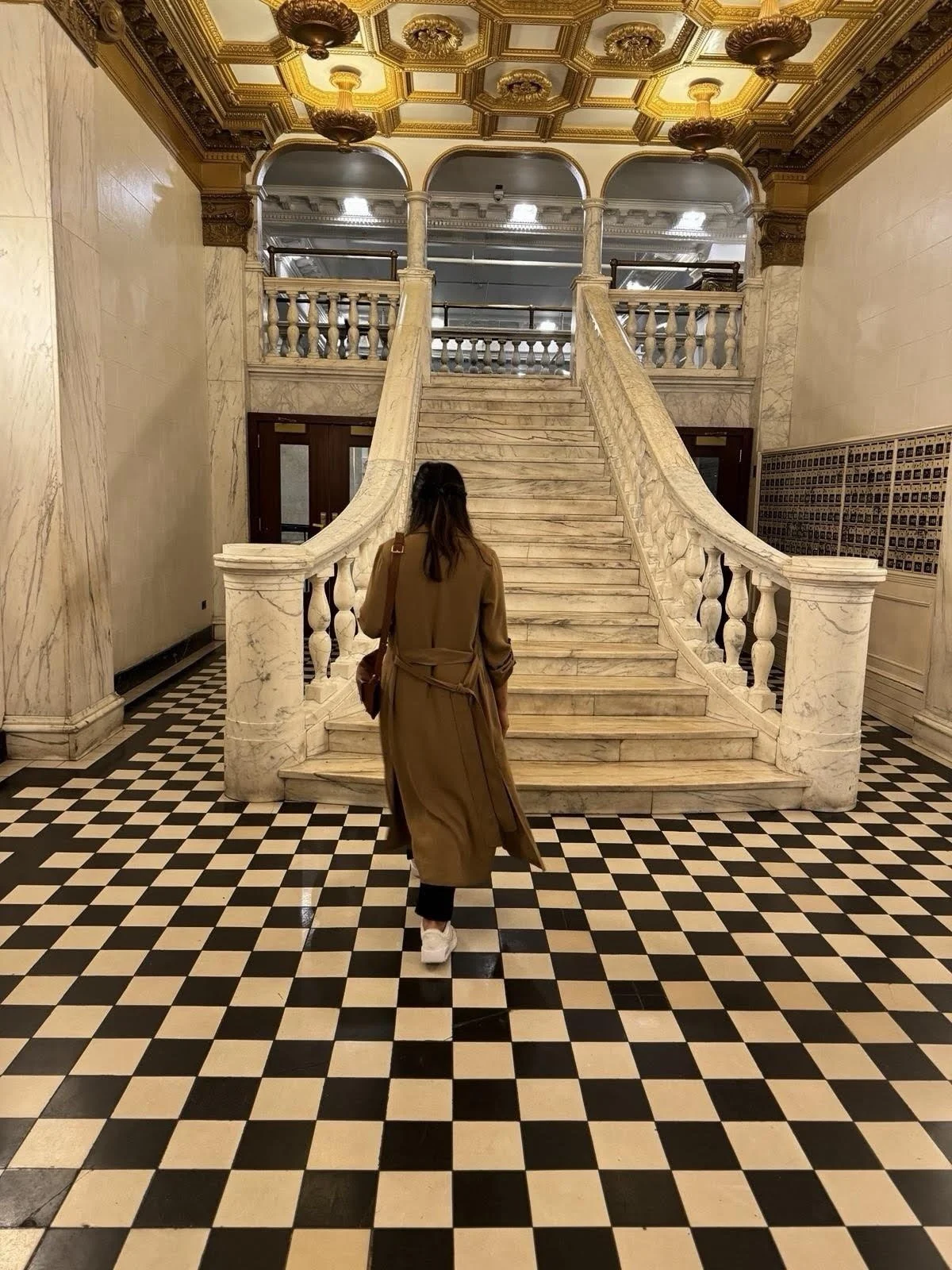 Woman walking towards a marble staircase inside a grand building with ornate gold ceiling decorations and checkered black and white floor tiles.