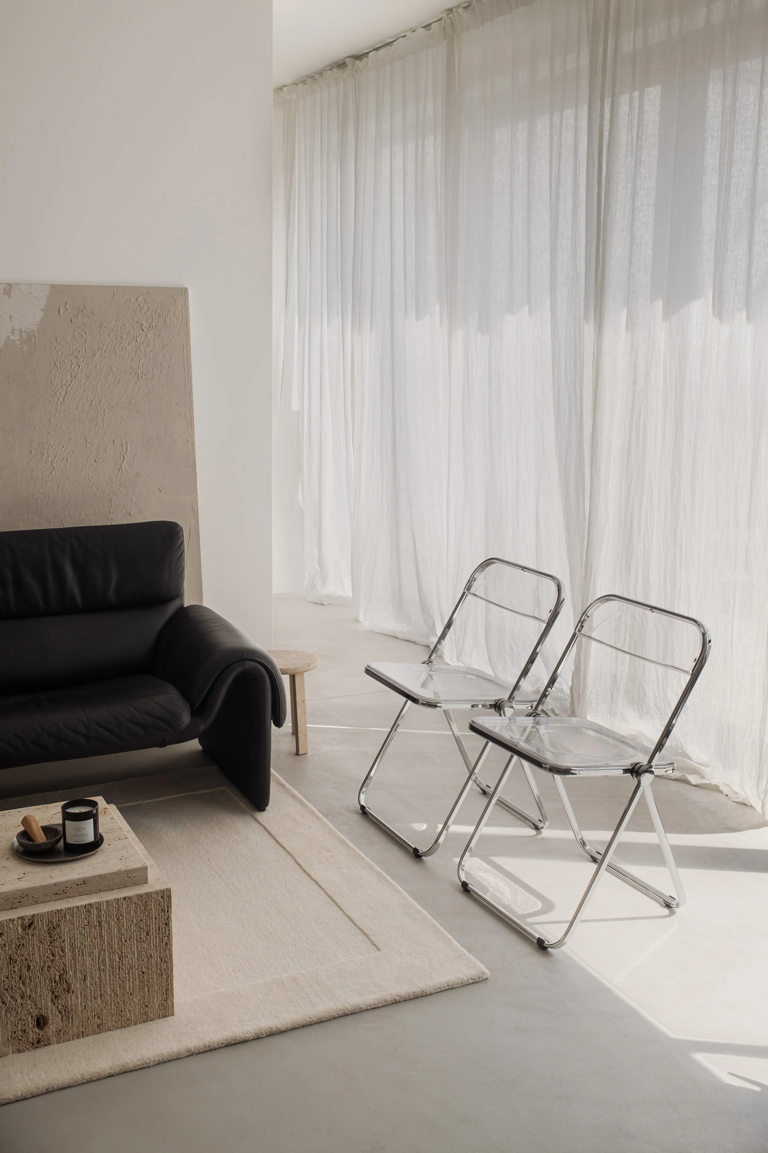 A modern, minimalist living room with white walls and sheer white curtains. There are two transparent chairs with metal frames, a black leather armchair, a small beige table, and a textured beige coffee table with a candle and tray on top. Light streams into the room, creating shadows on the floor.