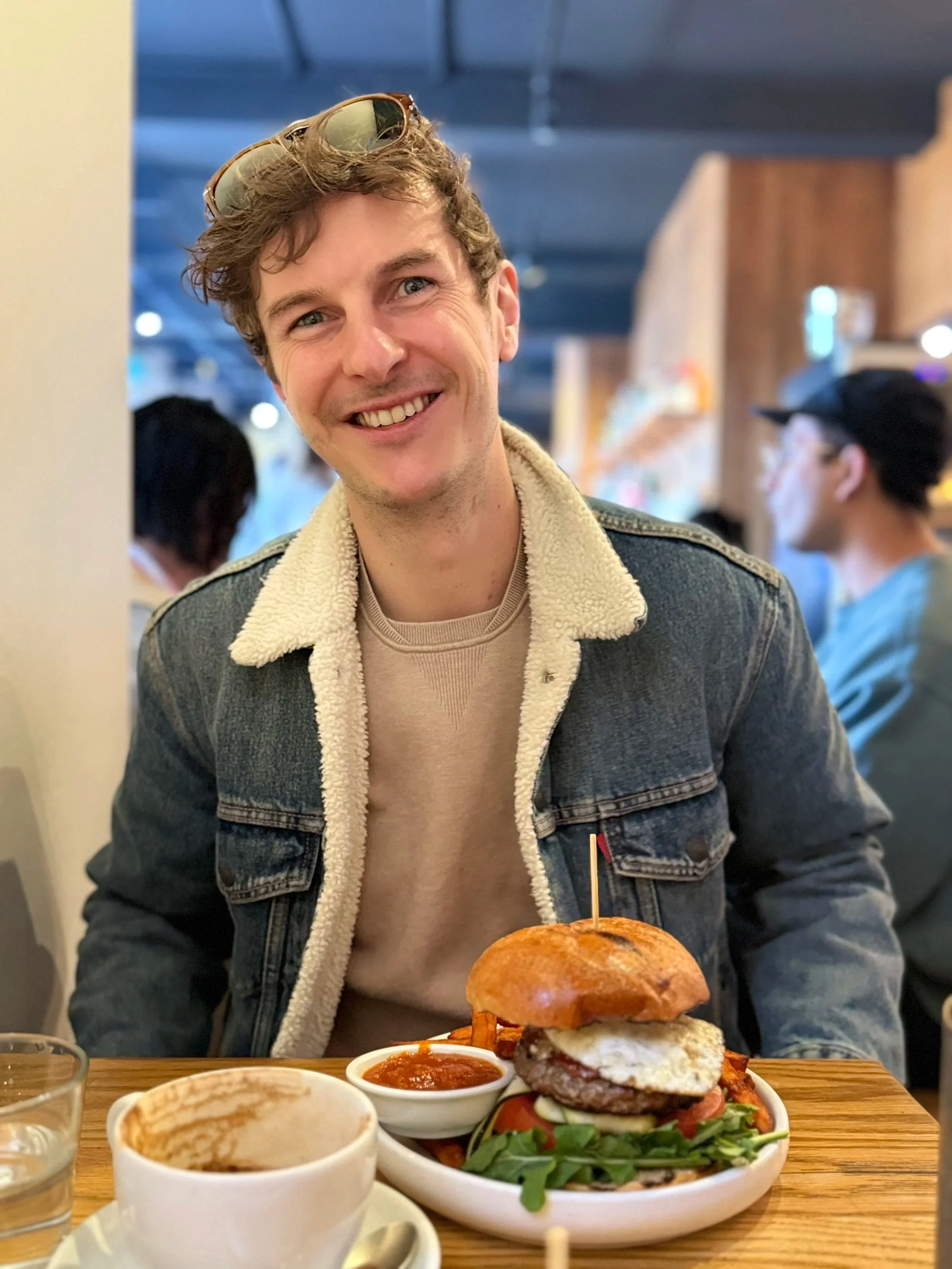 A portrait of poet Alex Scarborough. Taken in a restaurant with a huge burger in front of him. Alex is wearing a denim jacket and sunglasses on his head.