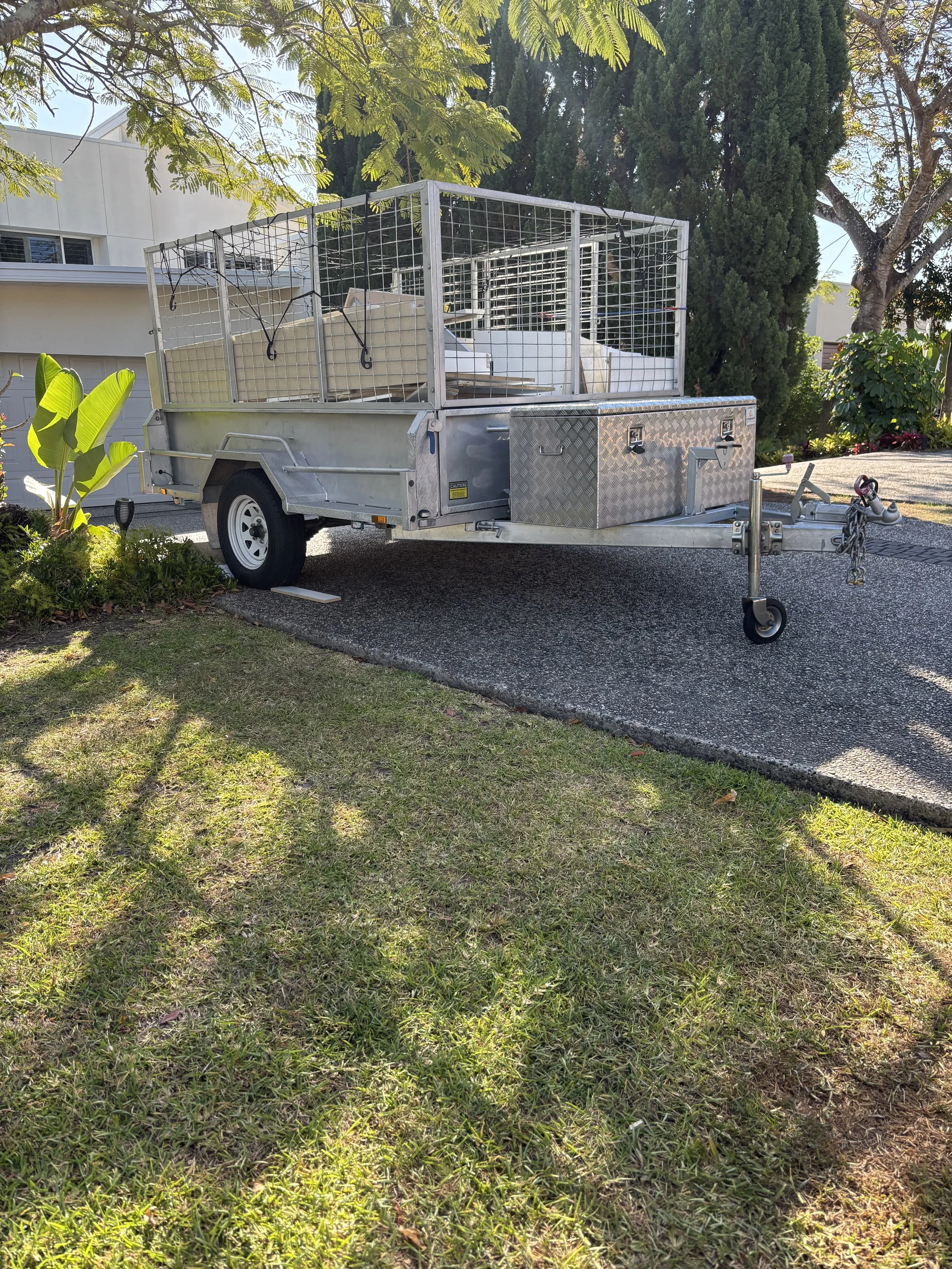 A small trailer with metal sides and a wire enclosure, parked on a driveway surrounded by grass and trees, with a white building in the background.