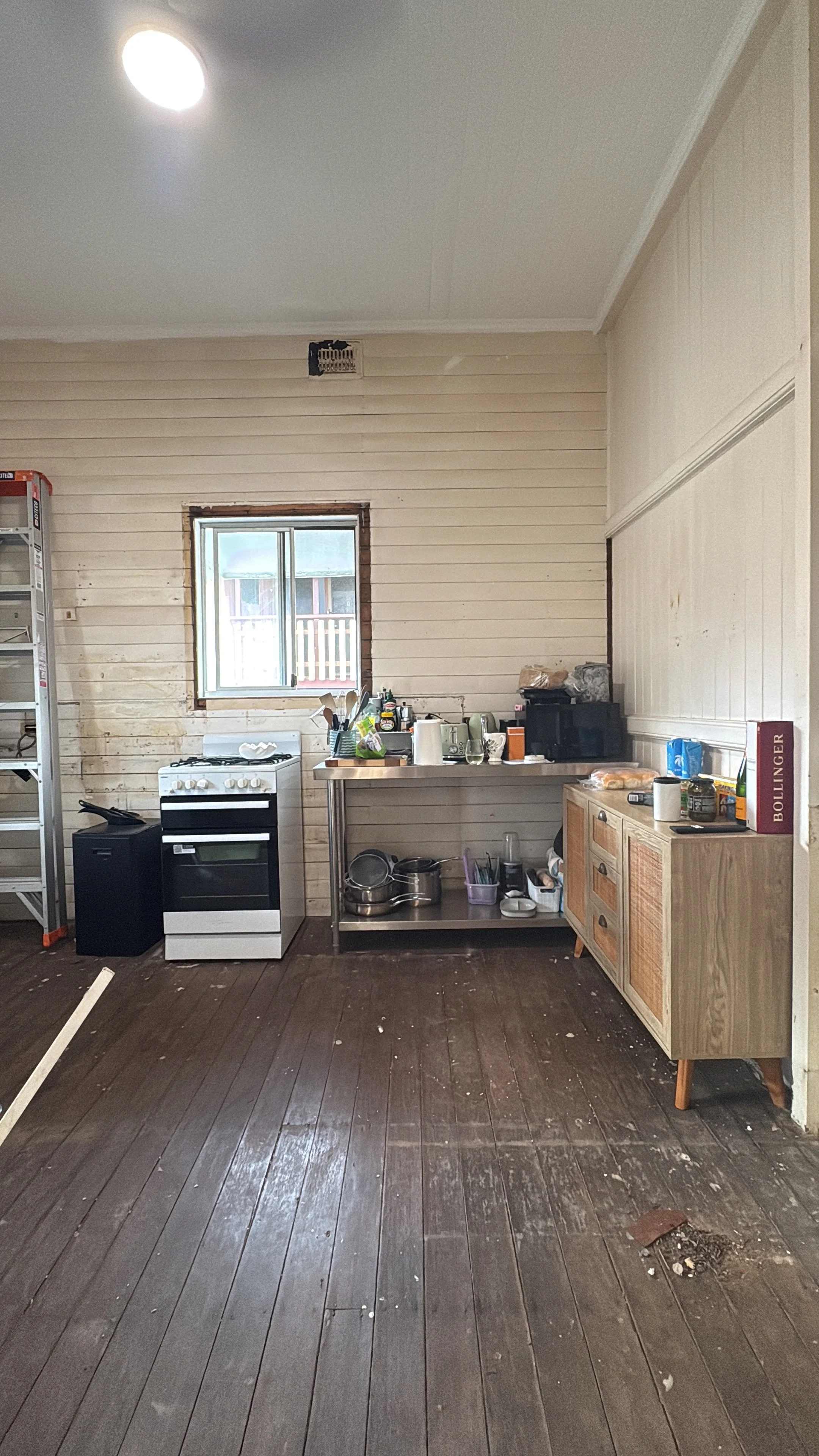 A rustic kitchen with wood paneling, a small window above a stainless steel countertop, and various kitchen appliances and utensils. The dark wooden floor is slightly dirty with debris.