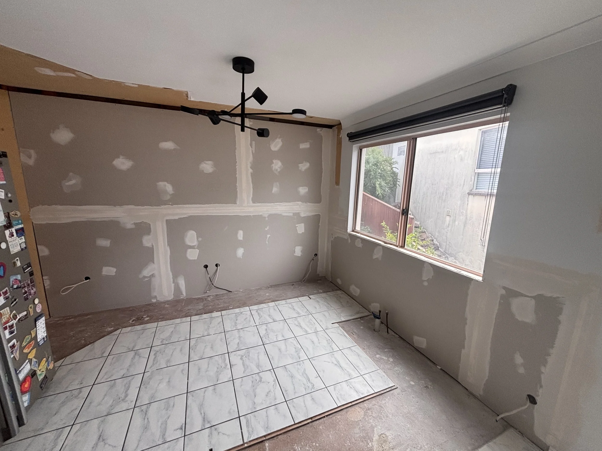 An unfinished kitchen with partially painted walls, a large window with blinds, and tiled floor. There is a modern black ceiling light fixture and visible wiring for appliances.