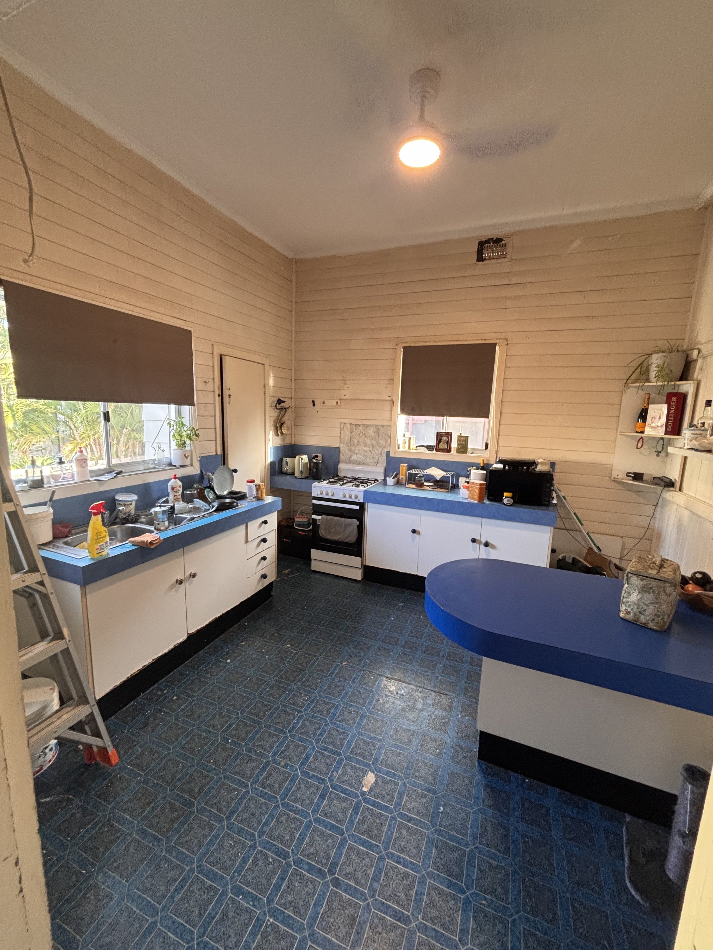 A cluttered kitchen with beige wooden paneling walls, blue and white cabinets, a gas stove, and a blue countertop. The window has a gray shade, and there are various household items on the counter and shelves, including bottles, a plant, and books. A