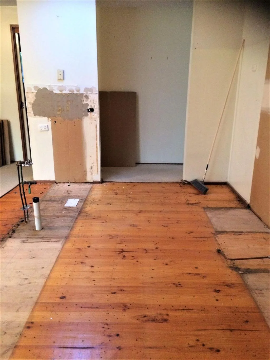 Room under renovation with unfinished walls, exposed pipes, a broom leaning against the wall, and wooden flooring partially installed.