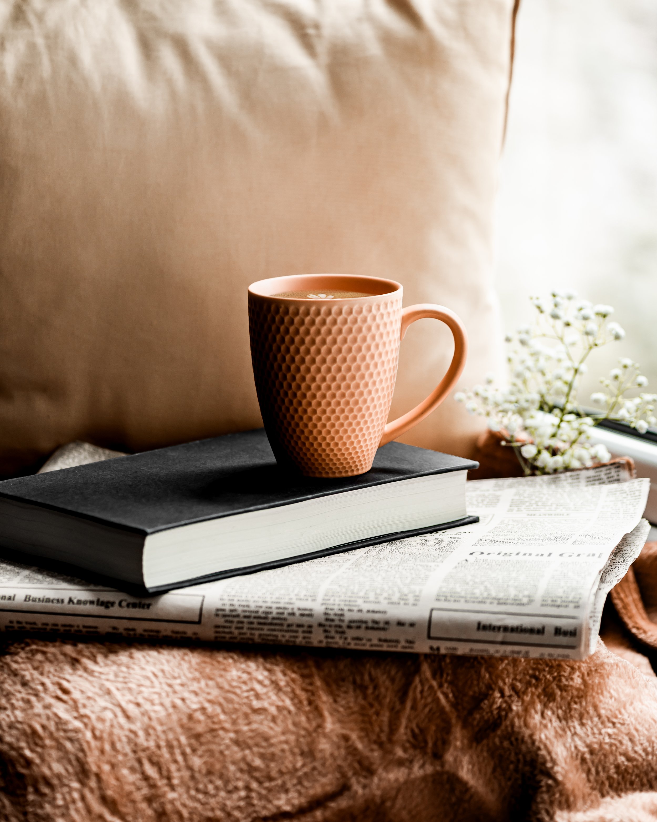 A beige ceramic mug with a textured honeycomb pattern, filled with a hot beverage, sitting on top of two closed books, with a black cover book on top. The scene includes a window near the right, with white flowers in a small vase on the windowsill, and a beige textured wall in the background.